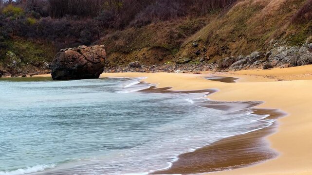 Large rock formation on a sandy beach in Spain. Coastal landscape featuring a boulder in the sea, golden sand, and a scrubby hillside background.