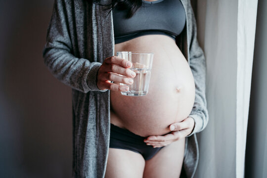 Pregnant woman holding drinking glass while touching belly at home