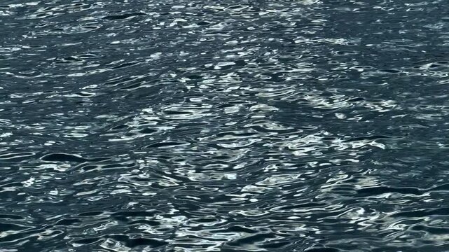 Rippled dark blue water surface with silver sunlight reflections. High angle view of textured water ripples with shimmering light glints and deep shadows.