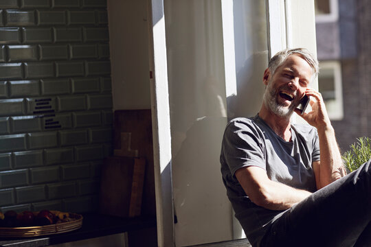 Laughing mature man sitting at the window at home talking on the phone