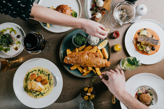 Overhead view of a shared meal with fish and chips, assorted plated dishes, and hands reaching for food. Two people enjoy a warm, convivial dining experience together