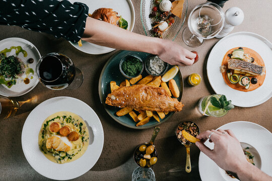 Overhead view of gourmet meal with fish, chips, and colorful side dishes. Hands reach for food, showing casual dining enjoyment. Plate arrangement suggests fine dining or celebratory gathering