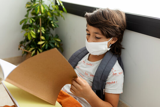 Close-up of boy wearing mask reading book while sitting against wall in school