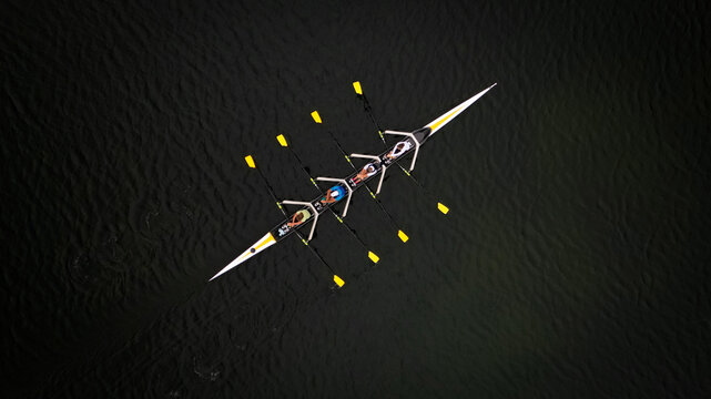 Aerial view of a sleek rowing boat cuts through the dark, still waters, its bright yellow oars a stark contrast to the shadowy depths, Divonne-les-Bains, Auvergne-Rhone-Alpes, France.