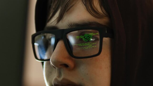 Teenage hacker in hood and glasses working at computer writing code. Program symbols reflecting in glasses lenses. Young programmer cyber criminal concept.