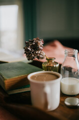 Warm cup of coffee sits beside milk bottle and dried hydrangea. Vintage book and small glass jar rest nearby. Soft light fills cozy, calm morning scene. Perfect for blogs, social media