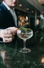 Man garnishes cocktail with herb in dimly lit bar. Hand places fresh green garnish over drink. Dark marble counter reflects glass and light