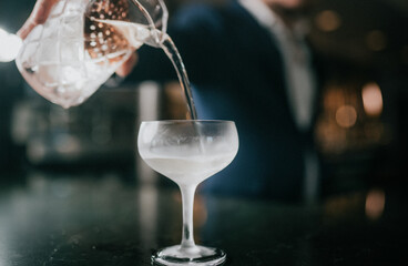 Cocktail glass being filled with clear liquid from jigger. Bartender pours drink with precision. Glass sits on dark bar surface. Background blurred with warm lighting. Perfect for ads, menus