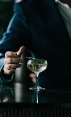 Bartender places a fresh mint leaf on a cocktail glass. Dark suit and gloved hand suggest elegance and precision. Glass holds clear liquid, possibly gin or champagne. Mood is sophisticated, calm © Reuben