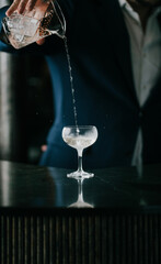 Hand pours clear liquid into cocktail glass. Bartender in suit prepares drink with care. Dark background highlights glass and pour. Perfect for bar, nightlife, or celebration themes