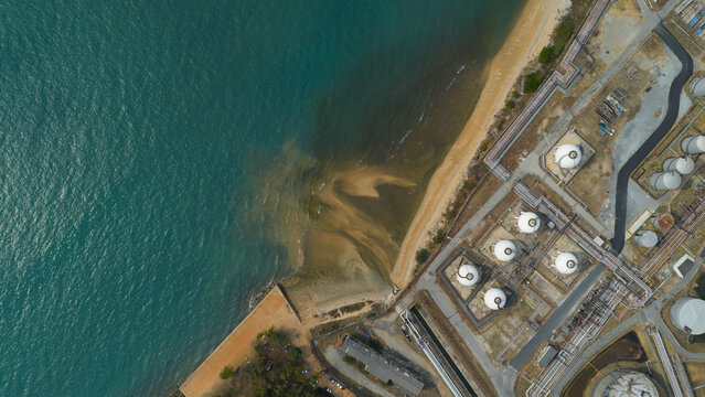 Aerial view of industrial plant with multiple tanks near the coastline where the dark turquoise ocean meets the light brown sand, Johor, Malaysia.