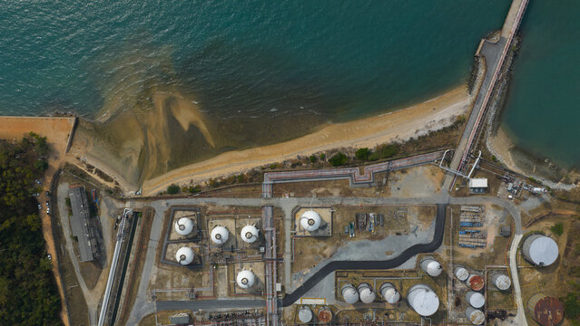 Aerial view of industrial tanks and pipelines meeting the turquoise sea, contrasting with the sandy beach, in Rayong, Thailand.