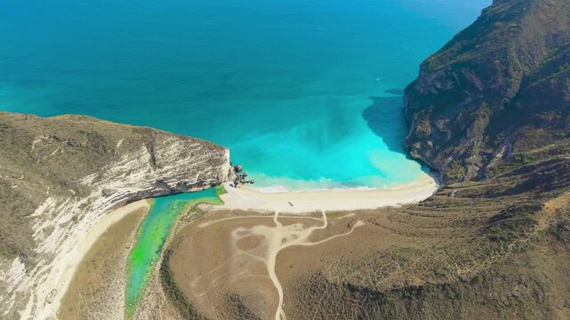 Aerial view of the turquoise water and white sand of Afoul Beach surrounded by steep rocky cliffs and desert terrain in Salalah, Oman.