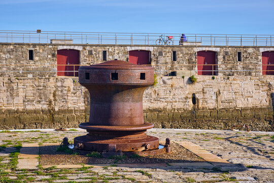 View of a weathered, rusted bollard standing sentinel against the backdrop of an aged stone wall with red-framed doors, Saint-Jean-de-Luz, France.