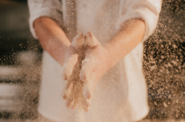 Fototapeta premium Close-up of hands catching falling flour dust. Person wears white apron, suggests baking or cooking. Warm, soft lighting highlights texture of flour particles. Ideal for recipes, food blogs