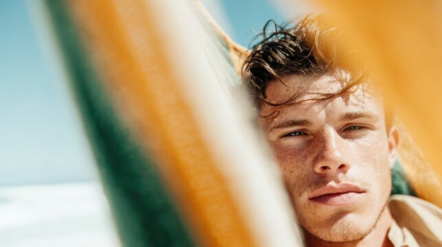 A handsome young man with tousled hair relaxes next to colorful fabrics by the beach, radiating a carefree vibe, connection to nature, and the warmth of summer days.