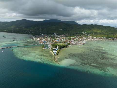 Aerial view of the vibrant town square meeting the tranquil turquoise sea, contrasting with the lush green hills under a cloudy sky, Karimunjawa, Central Java, Indonesia.