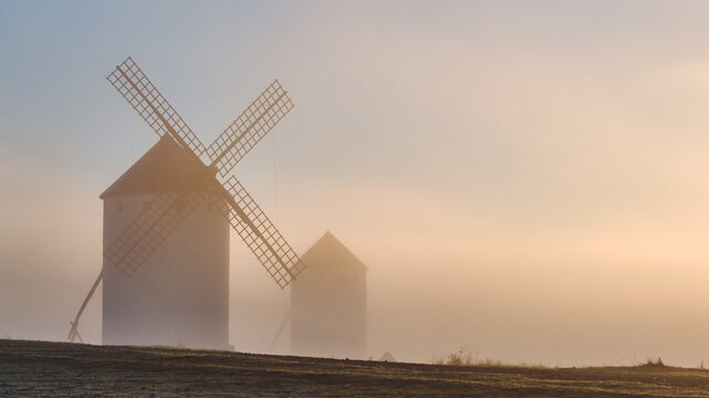 View of ancient windmills rising through the heavy fog, their sails silhouetted against the soft glow of dawn, Campo de Criptana, Castile-La Mancha, Spain.