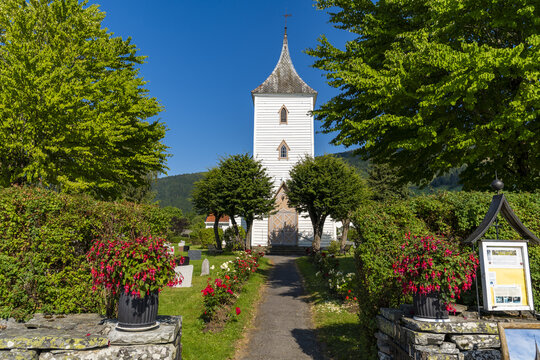 View of a pristine white Utvik church steeple, framed by vibrant green trees and bright red flowers, beckons down a stone path, Utvik, Vestland, Norway.