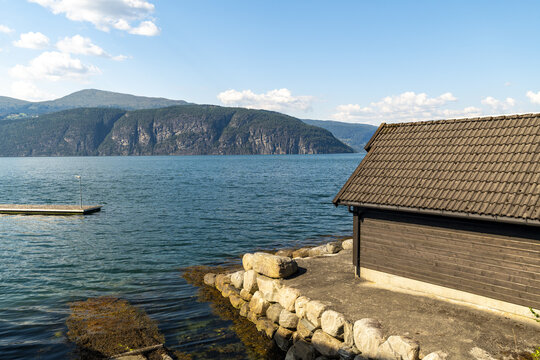 View of serene waters reflecting the clear sky, contrasted by the dark, textured mountains and a quaint boathouse, Utvik Fjord, Vestland, Norway.