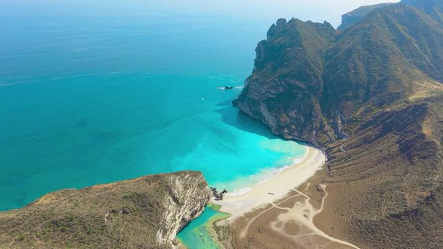 Aerial view of the turquoise water and white sand at Afoul Beach surrounded by steep coastal cliffs and mountains near Salalah, Oman.