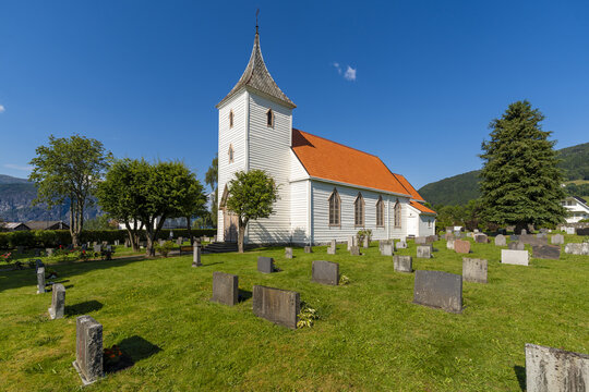 View of a quaint white Utvik church with an orange roof nestled among verdant trees and weathered gravestones under a clear blue sky, Utvik, Vestland, Norway.