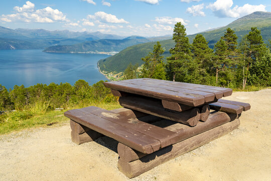 View of a rustic picnic table overlooks the serene Innvik fjord waters and distant mountains under a bright sky, Utvik, Vestland, Norway.