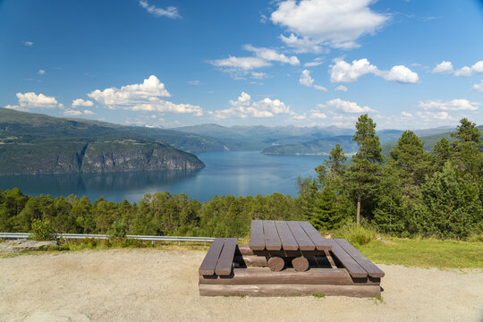 View of tranquil waters of Innvik Fjord mirroring the sky's azure canvas, framed by verdant hills and a rustic picnic table under the sun, Utvik, Vestland, Norway.