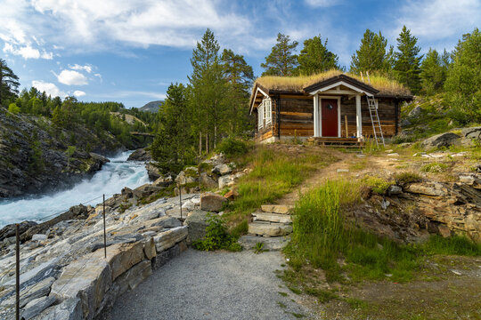 View of a quaint wooden cabin with a grass roof sits atop a hill overlooking a vibrant, rushing Tora river under a blue sky, Billingen, Norway.