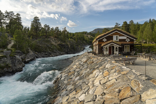 View of a cabin with a grass roof sits beside a rushing Tora river, the scene framed by lush greenery and a stone embankment, Billingen, Norway.