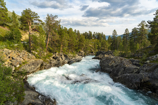 View of rushing turquoise Tora river cascading over dark, rugged rocks, flanked by verdant trees under a sky brushed with clouds, Billingen, Norway.