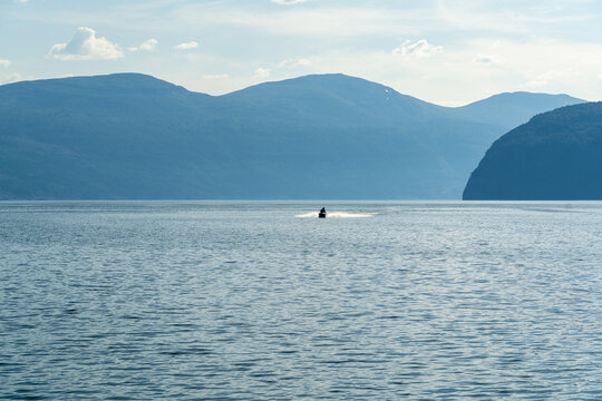 View of a lone jet ski cuts through the tranquil, sunny waters of the fjord, framed by layers of majestic, blue-tinged mountains, Innvik Fjord, Vestland, Norway.