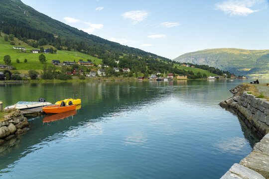 View of serene waters reflecting the lush green mountains and colorful boats docked near a stone pier under a clear sky, Olden, Vestland, Norway.