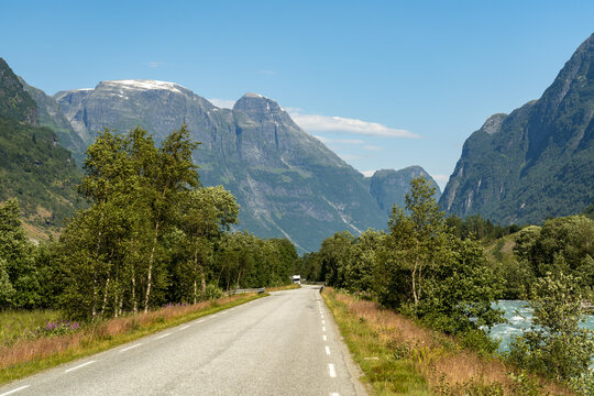 View of a long asphalt road winding through a verdant valley, framed by towering mountains under a clear blue sky, Oldevatnet, Vestland, Norway.
