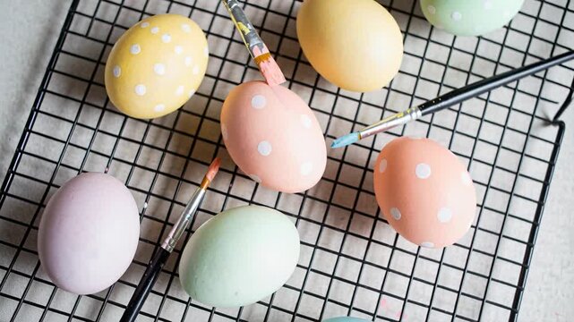 DIY paintbrushes shifting closer, applying white dots on pastel eggs on rack, creating patterns