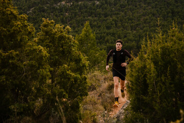 Fototapeta premium Male trail runner jogging along a narrow mountain path with scenic valley and countryside landscape in the background. Outdoor endurance sport and adventure lifestyle concept.