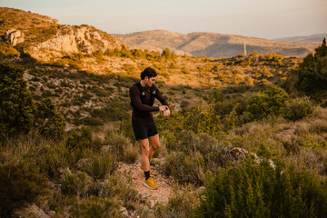 Fototapeta premium Young endurance athlete checking his smartwatch while training on a rugged mountain trail surrounded by natural landscape. Concept of trail running, outdoor fitness and endurance sport