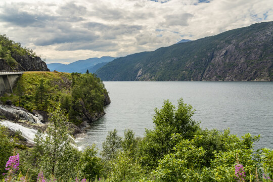 View of a serene Akra Glacial Fjord reflecting the sky, framed by verdant cliffs, a bridge, and wildflowers, creating a peaceful scene, Fjaera, Vestland, Norway.