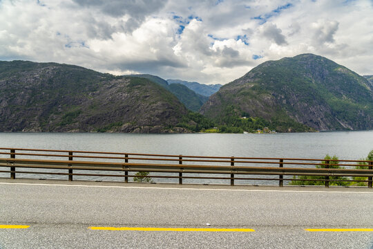 View of a serene lake reflecting the sky and mountains, framed by a bridge's simple railing and the road's yellow lines, Fjaera, Vestland, Norway.