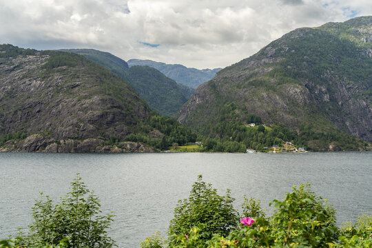 View of the calm lake reflecting the majestic, dark, rugged mountains under a cloudy sky, with a few houses nestled on the green shores, Akra Glacial Fjord, Fjaera, Vestland, Norway.