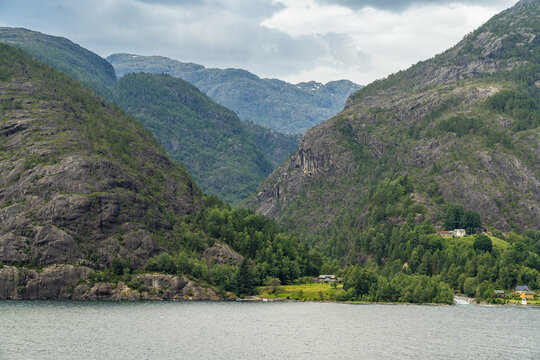 View of still waters reflecting the gray sky and the dramatic, steep, rocky mountains covered with lush green trees, Fjaera, Vestland, Norway.