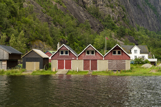 View of boathouses with red doors lining a calm waterfront against a backdrop of lush green trees and a rugged mountain, Fjaera, Vestland, Norway.