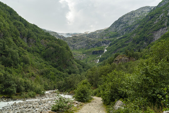 View of lush green mountains embracing a path alongside a rocky river leading to a distant glacier, a serene vista, buarbreen, Odda, Norway.
