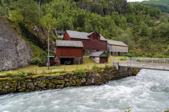 View of rustic red buildings nestled against a mossy cliffside, with a roaring river flowing under a modern bridge, set against a lush forest backdrop, buarbreen, Norway.