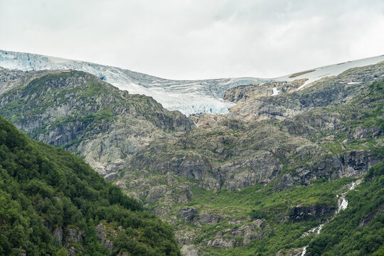 View of rugged, rocky mountainsides, splashed with verdant forests, rise to meet a glacial expanse under a soft, overcast sky, Buarbreen, Odda, Norway.
