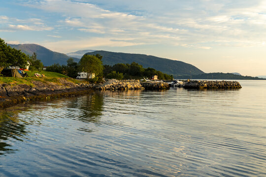 View of serene waters reflecting the sky, meeting a rocky shore dotted with green trees and mountains in the distance, Hardanger Fjord, Rosendal, Vestland, Norway.