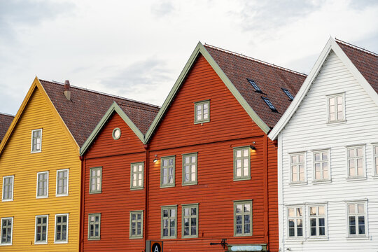 View of vibrant wooden buildings with striking gabled roofs stand shoulder-to-shoulder, a kaleidoscope of yellow, orange, and white hues against a cloudy sky, Bryggen, Bergen, Norway.