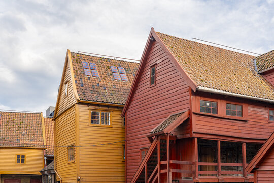 View of vibrant red, yellow, and brown painted wooden buildings with tiled roofs stand boldly against a cloudy sky, Bryggen, Bergen, Norway.