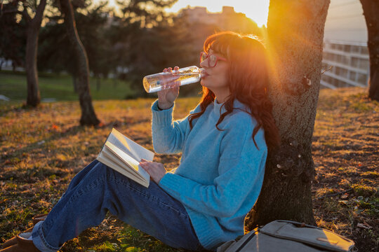 Woman drinking water and sitting with book near tree trunk at sunny day