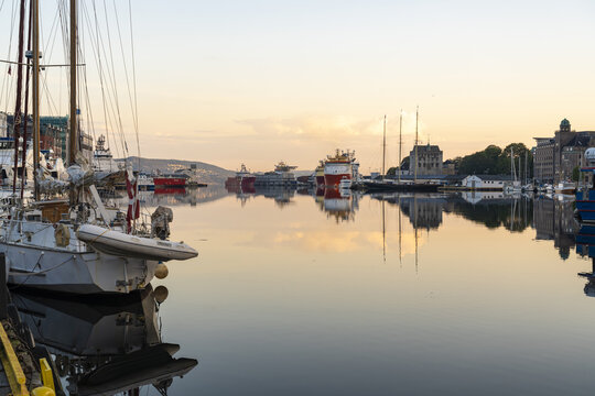 View of serene waters reflecting the golden sunrise over the harbor filled with various boats and ships in Bergen, Vestland, Norway.